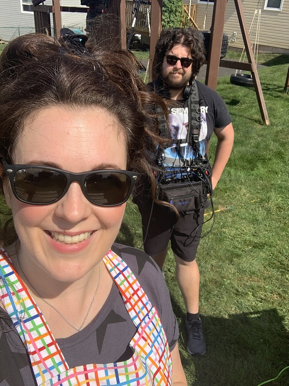 Heather Laura Clarke wearing a painting apron and sunglasses, posing in front of a camera operator during a TV shoot.